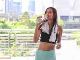 healthy and attractive Asian sportswoman in sportswear standing with  bridge fence and city building background, drinking water. photo