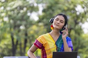 Asian transgender LGBTQ woman is listening to music from laptop while sitting relaxingly in the public park during summer with copy space photo