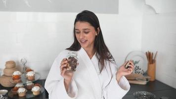 Young woman in robe holds jars of seasoning or food in bright white kitchen walks and talks toward camera video