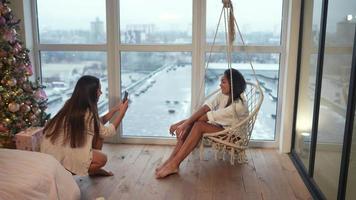 Young woman sits and swings in a hanging macrame chair by a window as friend photographs video