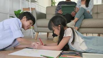 Asian Thai siblings are lying on living room floor, drawing homework with colored pencils together, parents leisurely relax on a sofa, lovely happy weekend activity, and domestic wellbeing lifestyle. video