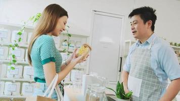 Asian male shopkeeper describes natural organic products to woman customer in refill store, zero-waste and plastic-free grocery, eco environment-friendly, sustainable lifestyles with a reusable shop.