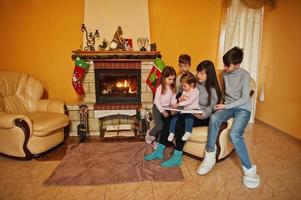 familia numerosa joven feliz junto a una chimenea en la cálida sala de estar en el día de invierno. madre con cuatro hijos en casa lee el libro. foto