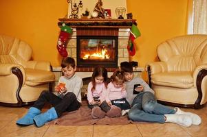 Happy four kids watching in they mobile phone at home by a fireplace in warm living room on winter day. photo