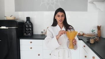 Young woman in robe holds jar of dry pasta in bright white kitchen walks and talks toward camera video