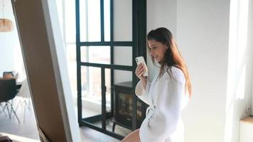 Young woman in robe video chats on smart phone standing before a mirror