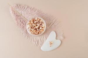 A face serum in pink capsules in the form of heart and a white Guasha face massager lying on a pink background photo