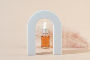 A dropper bottle with an orange nail cuticle oil standing in an arch in front of the pink feather on a beige background photo