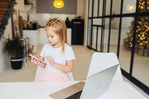 un niño pequeño se sienta en el escritorio con una computadora portátil y sostiene un teléfono móvil foto
