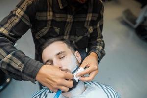 Young bearded man getting shaved by hairdresser at barbershop photo