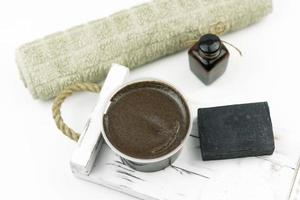 A chocolate scrub for body, a black hand made soap, a cooton towel, a natural oil for face stand on the white wooden tray in a modern bathroom photo