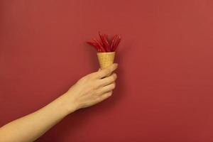 A womans hand holds mini red hot peppers in the waffle cone on a red background photo