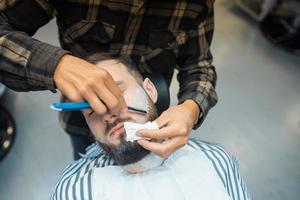 Young bearded man getting shaved by hairdresser at barbershop photo