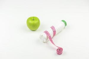 A green apple and a bottle of yogurt with a measuring tape on it lie on a white table. Measuring weight and weightloss concept photo