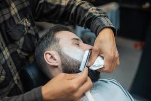 Young bearded man getting shaved by hairdresser at barbershop photo