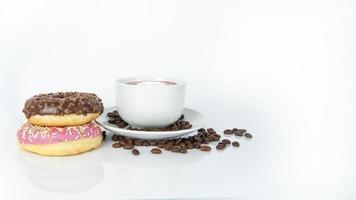 Aromatic coffee latte with leaf on the top in white cup on plate covered by brown seeds next to doughnuts. Chocolate and raspberry snacks on white background. Isolated objects in composition photo