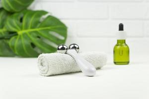 un aceite verde cbd, un rodillo para masaje facial, una toalla de algodón blanco y hojas verdes de monstera sobre una mesa blanca en un baño foto