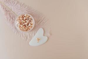 A face serum in pink capsules and a white Guasha face massager in the form of heart lying on a pink background photo