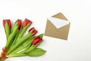 Red tulips and a blank letter in a brown envelope lie on a white table. Mockup photo