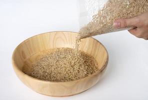 Woman's hand is pouring raw brown rice packed in a plastic bag onto a wooden bowl, on a white background. photo