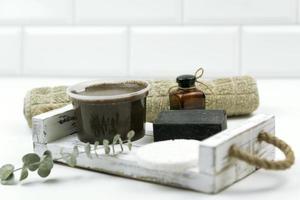 A chocolate scrub for body, a black hand made soap, a cooton towel, a white sponge and natural oil for face stand on the white wooden tray in a modern bathroom photo