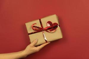 A womans hand holds a gift box with red heart and ribbon on it photo