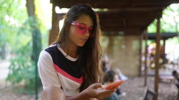 jovem mulher em óculos de sol rosa textos no telefone inteligente e olha para a câmera para sorrir video