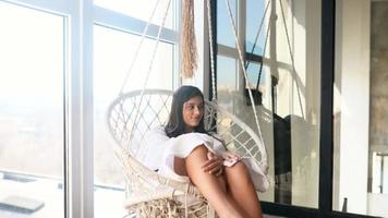 Young woman sits and swings in a hanging macrame chair by a window video