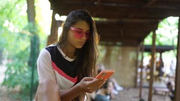 una joven con gafas de sol rosas escribe en un teléfono inteligente y mira a la cámara para sonreír video