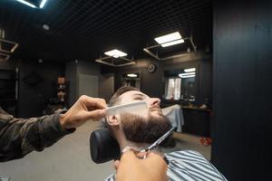 Hairdresser doing haircut of beard using comb and scissors photo