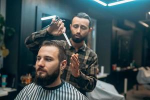 Cheerful young bearded man getting haircut by hairdresser at barbershop photo