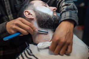 Young bearded man getting shaved by hairdresser at barbershop photo