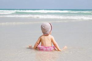 Little baby girl is sitting at sandy beach and looking at the blue sea with waves. photo