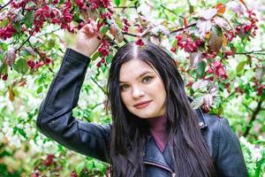 retrato de una mujer atractiva con el pelo largo y castaño con chaqueta de cuero en el jardín con flores de manzano rojo y blanco y gotas de lluvia. foto