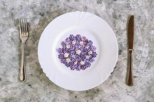 Served plate with purple lilac flowers with fork and knife on gray concrete background, top view. Fresh spring concept with minimal design.