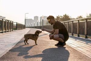 Young handsome man is feeding his dog from hand in the morning on empty street of city. photo