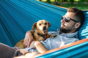 un apuesto hombre europeo con gafas de sol descansa en una hamaca con su lindo perrito. foto
