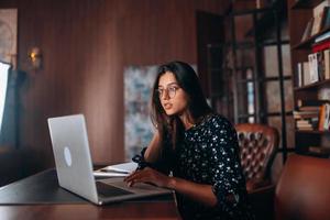 Young woman in glasses works at the laptop while sitting at table photo