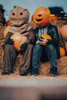 A guy with a  pumpkin head sits next to a scarecrow photo