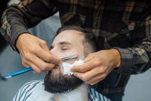 Young bearded man getting shaved by hairdresser at barbershop photo