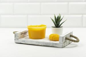 A yellow mango hand made soap, a mango scrub and a potted green succulent stand on a wooden tray in a modern bathroom photo