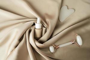 A face massage cream, a white Guasha face massager in the form of heart and face roller lying on a beige fabric photo