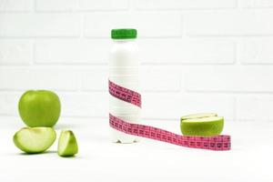 A green apple and a bottle of yogurt with a measuring tape on it stand on a white table. Measuring weight and weightloss concept photo