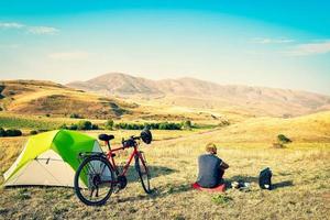 Caucasian man sit by touring bicycle in mountains on side of green tent have break time rest in morning. Solo travel journey. Long-term travel around the world photo