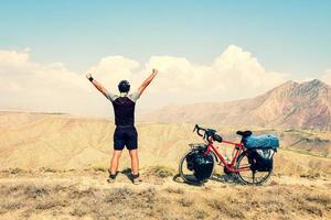 cinemático inspirador emocionado ciclista masculino caucásico parado en el punto de vista superior en bicicleta de gira roja en el fondo de las montañas desiertas con las manos en alto estilo de vida seguro y despreocupado foto