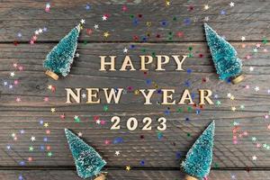 Happy New year 2023 celebration. Wooden text and numbers 2023 on wooden background with scattered multicolored confetti and fur tree miniature toys. photo