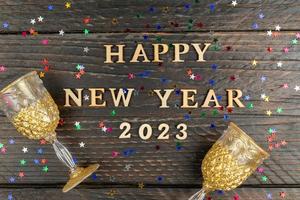 wooden text happy new year 2023 on wooden background surrounded multicolored stars confetti anf two glasses for champagne. Festive greeting card. photo