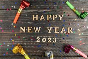 feliz año nuevo 2023 letras de madera sobre fondo de madera rodeado de silbatos multicolores de fiesta. tarjeta de felicitación festiva para las vacaciones de año nuevo foto