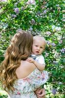 joven madre con un hijo pequeño en sus manos en el parque con un árbol de flor lila, vista trasera. feliz madre e hijo. foto