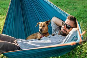 un joven europeo apuesto con gafas de sol descansa en una hamaca con su lindo perrito. foto